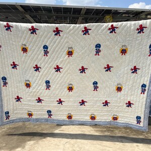 May include: A cream-colored quilted blanket featuring a repeating pattern of embroidered superhero figures, including Spider-Man and Captain America. The blanket is finished with a blue and white chevron-patterned border along all four edges.