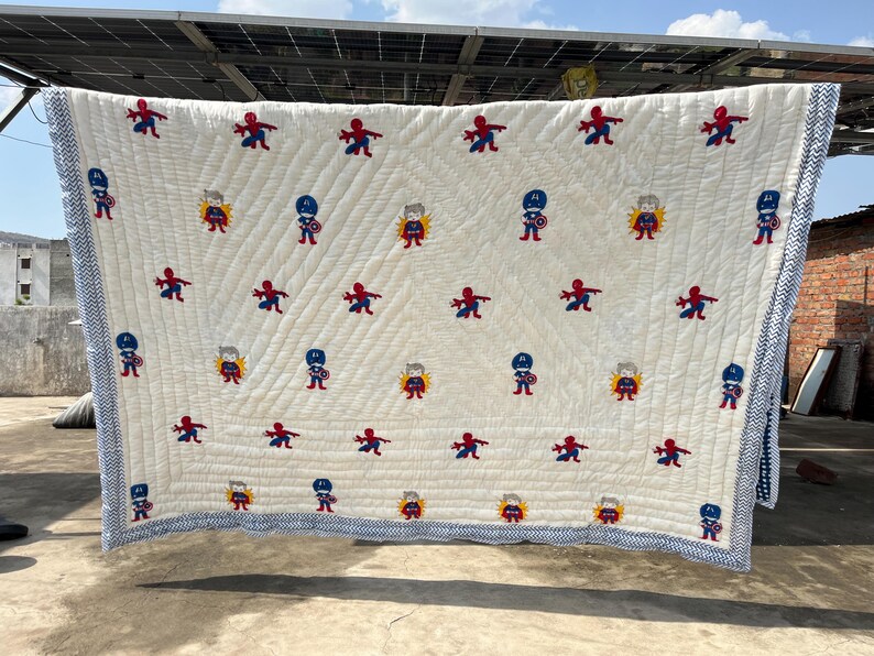 May include: A cream-colored quilted blanket featuring a repeating pattern of embroidered superhero figures, including Spider-Man and Captain America. The blanket is finished with a blue and white chevron-patterned border along all four edges.