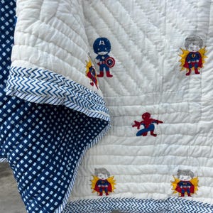 May include: A white quilted blanket featuring a repeating pattern of cartoon superhero characters including Captain America, Superman, and Spider-Man. The blanket has a dark blue and white checkered reverse side and a blue chevron patterned border trim.