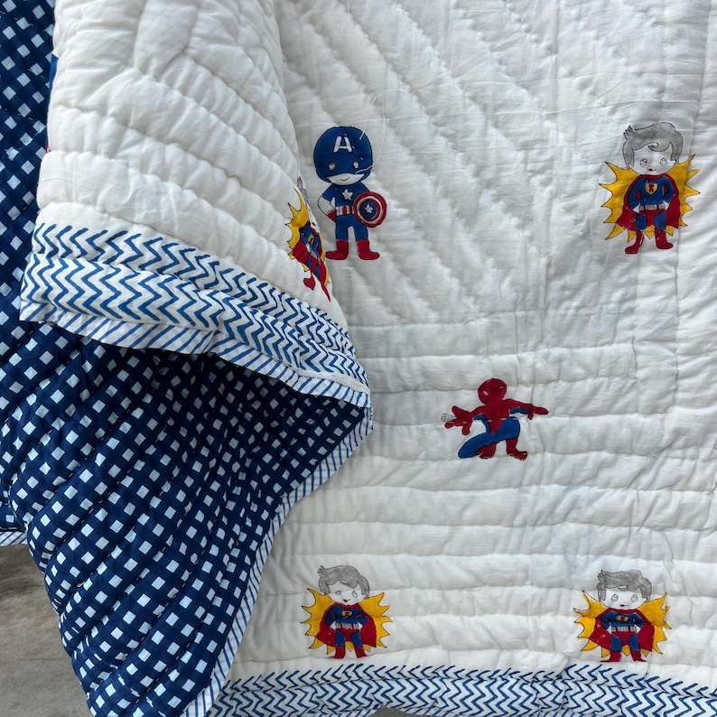 May include: A white quilted blanket featuring a repeating pattern of cartoon superhero characters including Captain America, Superman, and Spider-Man. The blanket has a dark blue and white checkered reverse side and a blue chevron patterned border trim.