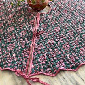 Puede incluir: Una falda de &aacute;rbol redonda y acolchada con un estampado floral rosa y verde. La falda tiene un borde festoneado y lazos rosas. El dise&ntilde;o floral se repite en toda la superficie, creando un aspecto decorativo y cohesivo.