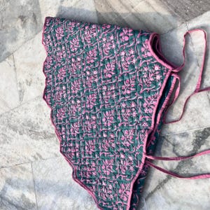 Puede incluir: Textil acolchado con borde festoneado, con un patr&oacute;n floral repetido en rosa sobre un fondo verde oscuro. Los bordes est&aacute;n ribeteados con tela rosa y se ven lazos rosas. El textil est&aacute; doblado.