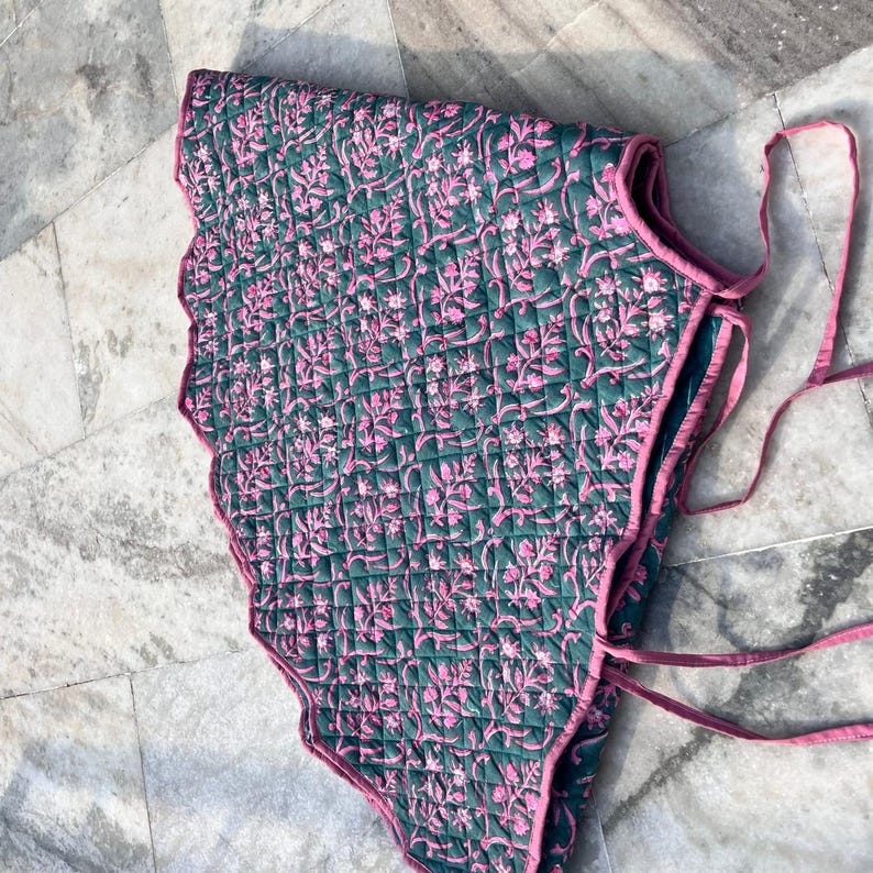 Puede incluir: Textil acolchado con borde festoneado, con un patr&oacute;n floral repetido en rosa sobre un fondo verde oscuro. Los bordes est&aacute;n ribeteados con tela rosa y se ven lazos rosas. El textil est&aacute; doblado.