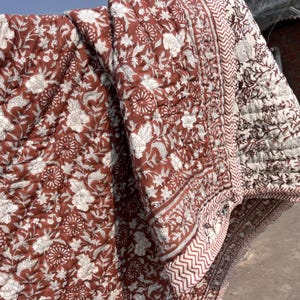 Puede incluir: Textil acolchado plegado con un diseño floral blanco sobre un fondo marrón rojizo. El patrón incluye flores y hojas. Los bordes están bordeados con un diseño geométrico. La construcción en capas del textil es visible.