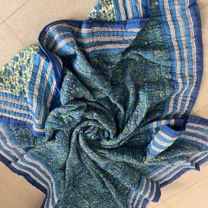 Könnte beinhalten: Eine blau-grüne Steppdecke mit floralem Muster und gestreiftem Rand. Die Steppdecke besteht aus weichem Stoff und weist ein detailliertes Blumendesign auf. Die Steppdecke ist gefaltet und drapiert und zeigt ihre Textur und ihr Muster.