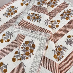 Könnte beinhalten: Eine gefaltete weiße Steppdecke mit einem floralen Muster in Gold- und Grautönen. Braune Streifenabschnitte wechseln sich mit den floralen Designs ab. Die Kanten sind mit einem grauen Zickzackmuster versehen.
