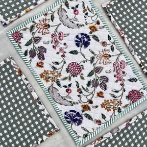 Può includere: Una tovaglietta trapuntata rettangolare con sfondo bianco e un motivo floreale multicolore nelle tonalità del viola, rosa, arancione e verde. La tovaglietta è rifinita con un sottile bordo in tessuto a righe verdi e bianche.