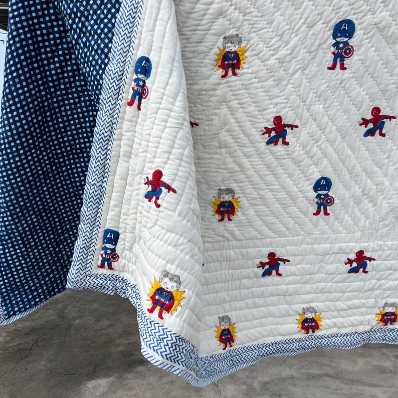 May include: A quilted blanket featuring a white center panel with embroidered superhero characters, including figures in red, blue, and yellow costumes. The blanket has a blue and white chevron border and a dark blue and white gingham side panel.