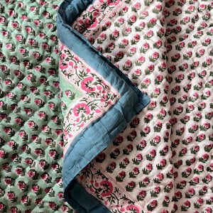 Könnte beinhalten: Eine florale Steppdecke mit hellgrünem und weißem Hintergrund. Die Steppdecke weist ein sich wiederholendes Muster aus rosa Blüten und grünen Blättern auf. Der Rand ist türkisblau mit einem floralen Design. Die Steppdecke ist gefaltet und zeigt das Muster im Inneren.