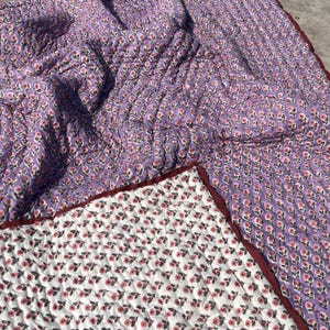 May include: A quilted blanket with a floral pattern. The top side is a lavender color with a repeating pattern of small pink flowers and green leaves. The underside is white with a similar floral design. The edges are trimmed with a dark red border.
