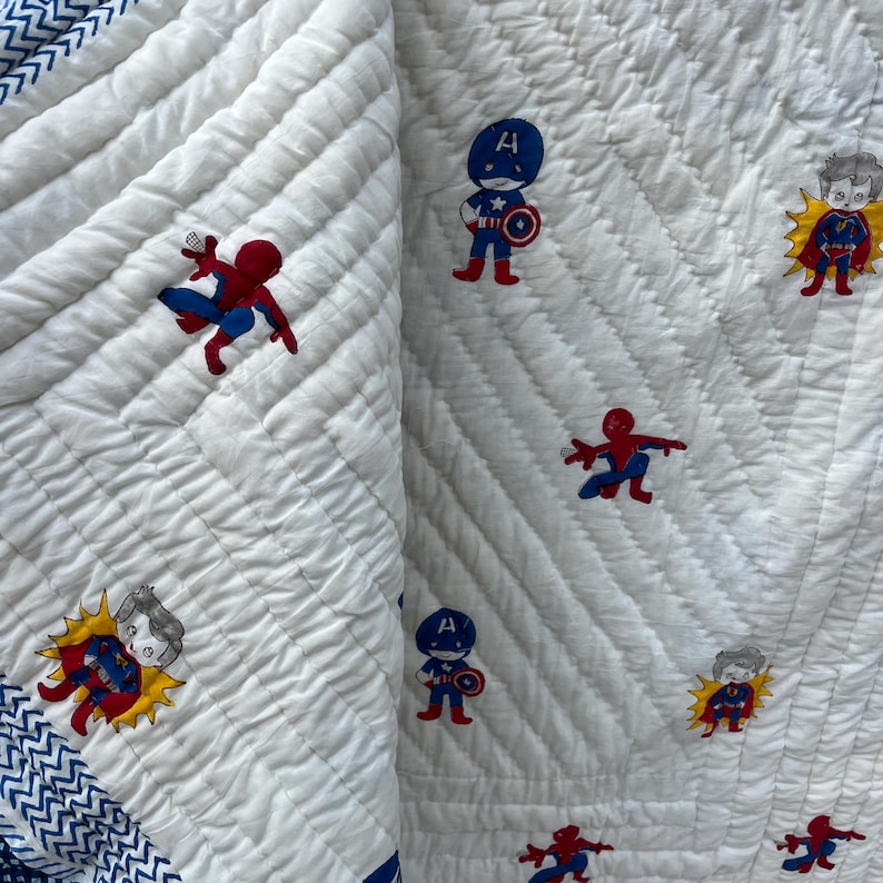May include: A white quilted blanket featuring a repeating pattern of cartoon superheroes including Spider-Man, Captain America, and Superman. The fabric has a textured, stitched surface and a blue and white chevron patterned border along the edge.