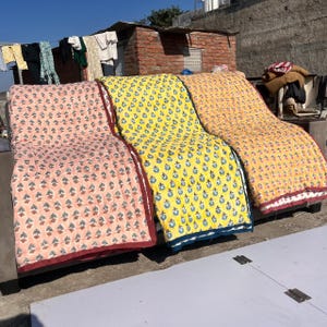 May include: Three colorful, patterned quilts are displayed outdoors. The quilts are in shades of pink, yellow, and orange, each with a different floral design. The quilts have a quilted texture and are bordered with contrasting colors. The background shows a brick building and clothes hanging on a line.