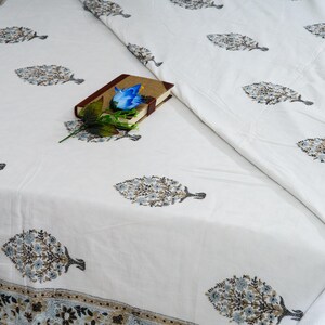 Könnte beinhalten: Weißes Tischtuch mit einem sich wiederholenden Muster aus stilisierten baumartigen Designs in Blau-, Grau- und Brauntönen. Ein kleines braunes Buch mit einer blauen Blume und grünen Blättern liegt auf dem Tischtuch. Das Tischtuch hat einen dekorativen Rand.