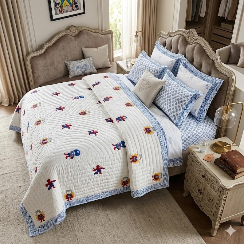 May include: A white quilted bedspread featuring a repeating pattern of embroidered superhero figures in red, blue, and yellow. The bedding set includes matching pillowcases with blue and white geometric borders and a central beige accent pillow.