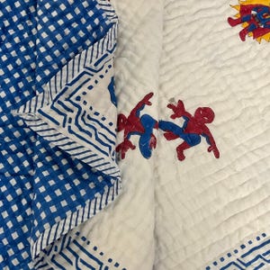 Könnte beinhalten: Eine weiß-blaue Steppdecke mit Superhelden-Motiv. Die Steppdecke hat auf der einen Seite ein blau-weißes Karomuster und auf der anderen Seite applizierte Superheldenfiguren in Rot und Blau. Die Steppdecke ist gefaltet und zeigt beide Seiten.