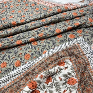 Könnte beinhalten: Eine Steppdecke mit floralem Muster mit orangefarbenen Blumen und grauen Blättern auf grauem Hintergrund. Die Steppdecke hat einen weiß-grauen Zickzack-Rand und eine weiße Ecke mit floralem Muster. Die Steppdecke ist gefaltet und scheint aus einem weichen Material zu bestehen.