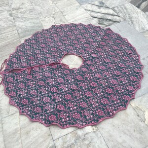 Puede incluir: Una falda de &aacute;rbol redonda con un borde festoneado. La falda es de color verde oscuro con un patr&oacute;n floral rosa repetido. Un ribete rosa bordea el borde y la abertura para el tronco del &aacute;rbol. Se adjuntan lazos para asegurar la falda.