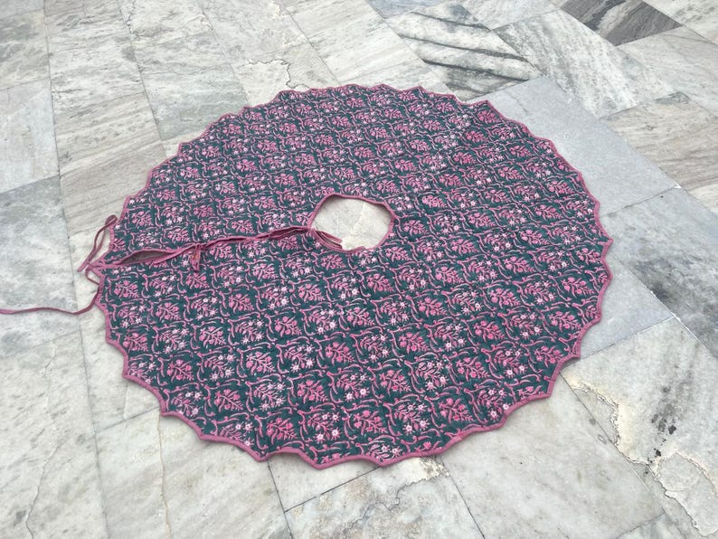 Puede incluir: Una falda de &aacute;rbol redonda con un borde festoneado. La falda es de color verde oscuro con un patr&oacute;n floral rosa repetido. Un ribete rosa bordea el borde y la abertura para el tronco del &aacute;rbol. Se adjuntan lazos para asegurar la falda.