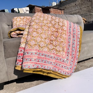 May include: A patterned quilt with a floral design in red, yellow, and white, resting on a gray sofa. The quilt has a red and gray border and a yellow backing. The quilt is folded and draped over the sofa.