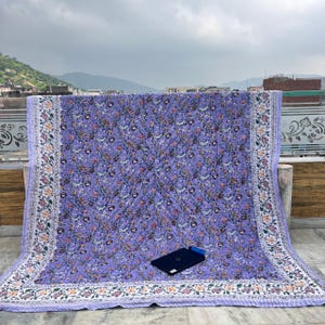 May include: A large, lavender floral quilt with a detailed border. The quilt features a repeating pattern of flowers in shades of pink, orange, and green. The border is white with floral and leaf designs. A laptop is placed on the quilt.