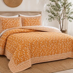 Könnte beinhalten: Eine gesteppte Tagesdecke und zwei passende Kissenbezüge in leuchtendem Orange mit einem weißen Blumenmuster. Das Bettwäscheset verfügt über eine dekorative Bordüre mit komplexen geometrischen und botanischen Motiven an den Rändern.