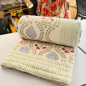 Könnte beinhalten: Aufgerollte cremefarbene Steppdecke mit floralem Muster. Die Steppdecke zeigt ein sich wiederholendes Design aus blauen und rosa Blumenmotiven und grünen Ranken auf cremefarbenem Hintergrund. Der Rand hat ein grünes Zickzackmuster.