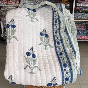 Könnte beinhalten: Eine weiße Steppdecke mit einem sich wiederholenden Muster aus blauen Blumenmotiven mit grünen Stielen. Die Decke hat einen breiten Rand mit einem blauen Blumenrankenmuster und hellgrünen Streifen, abgerundet durch vertikale Steppnähte.