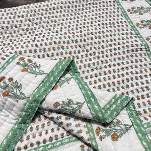 Könnte beinhalten: Eine weiße Steppdecke mit einem sich wiederholenden Muster aus kleinen orangefarbenen Blumen und grünen Stielen. Die Decke hat einen breiten Rand, der mit größeren orangefarbenen Blumenzweigen und grünen Blättern verziert ist, mit sichtbaren Nähten.
