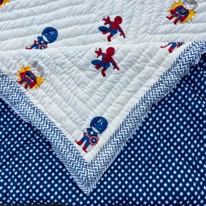 May include: A quilted blanket featuring a white top layer with a chevron-stitched texture and a pattern of cartoon superheroes including Spider-Man and Captain America. The underside is a dark blue fabric with a white grid pattern.