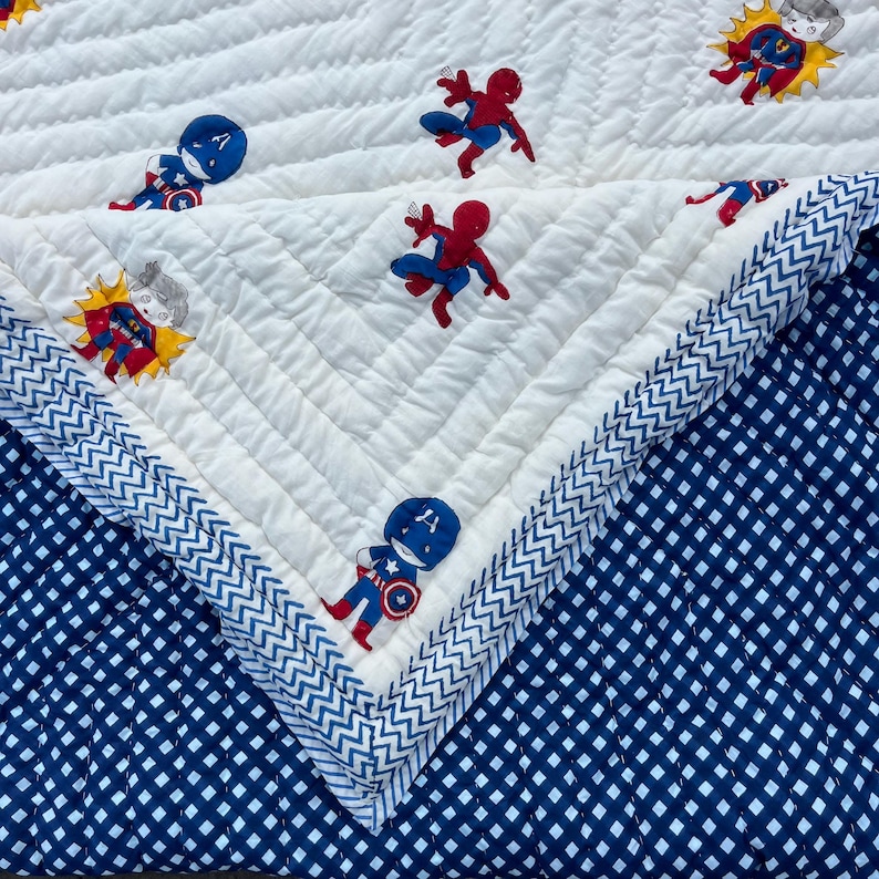 May include: A quilted blanket featuring a white top layer with a chevron-stitched texture and a pattern of cartoon superheroes including Spider-Man and Captain America. The underside is a dark blue fabric with a white grid pattern.