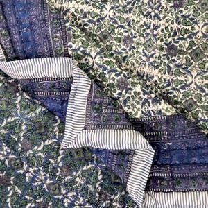 May include: A quilted textile featuring a floral pattern in shades of blue, green, and purple on a white background. The edges are trimmed with a striped fabric in white and blue. The quilt is folded, showcasing its design.