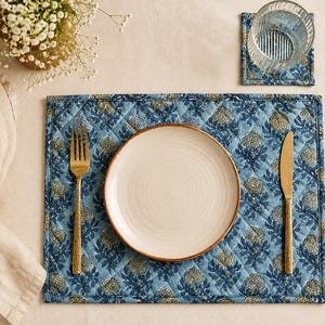 Könnte beinhalten: Ein gestepptes, rechteckiges Platzset in Hellblau mit einem sich wiederholenden dunkelblauen und beigen Blumenmuster. In der Mitte des Platzsets steht ein runder, cremefarbener Teller, flankiert von einer goldfarbenen Gabel links und einem Messer rechts.