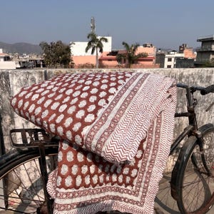 Peut inclure: Une couverture pliée, à motifs marron et blanc, est posée sur un vélo vintage. La couverture présente un motif floral répétitif et une bordure décorative. L'arrière-plan montre une scène extérieure ensoleillée avec des bâtiments et des arbres.