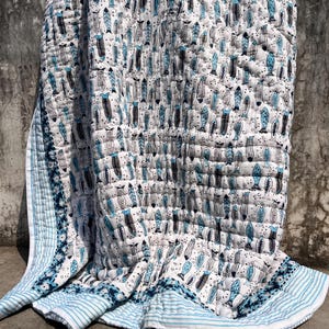 Könnte beinhalten: Eine weiße Steppdecke mit einem sich wiederholenden Muster aus kleinen Fischen in Blau- und Schwarztönen. Die Decke hat eine gesteppte Textur und ist am unteren Rand mit einer blau-weiß gestreiften Bordüre versehen.