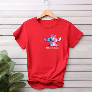 May include: A red t-shirt featuring a graphic of two cartoon characters embracing, one blue and one pink. The text "Robert & Jessica" is printed below the graphic. The t-shirt is hanging on a wooden hanger.