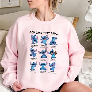 Op de afbeelding: Roze sweatshirt met de tekst "GOD SAYS THAT I AM..." en afbeeldingen van een blauw stripfiguur. Het personage wordt in verschillende poses getoond met woorden als "EMPOWERED", "TREASURED" en "PRECIOUS".