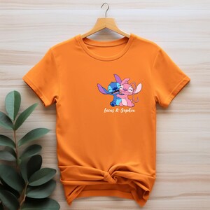 May include: Orange t-shirt featuring a cartoon illustration of two characters embracing, one blue and one pink. The text "Lucas & Sophia" is printed below the characters. The t-shirt is hanging on a wooden hanger.