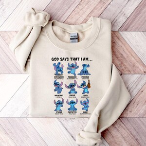 Op de afbeelding: Een cr&egrave;mekleurige sweatshirt met een afbeelding van het personage Stitch uit Lilo & Stitch. Het ontwerp bevat Stitch in verschillende poses met woorden als "Empowered", "Treasured" en "Precious". De tekst "GOD SAYS THAT I AM..." staat bovenaan.