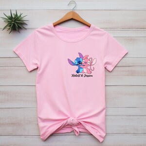 May include: A light pink t-shirt featuring a graphic of two cartoon characters embracing, with the text "Robert & Jessica" below. The characters are blue and pink. The shirt is knotted at the bottom.