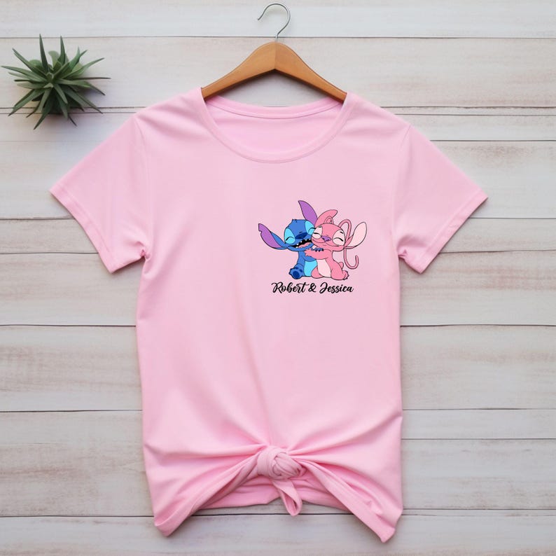 May include: A light pink t-shirt featuring a graphic of two cartoon characters embracing, with the text "Robert & Jessica" below. The characters are blue and pink. The shirt is knotted at the bottom.