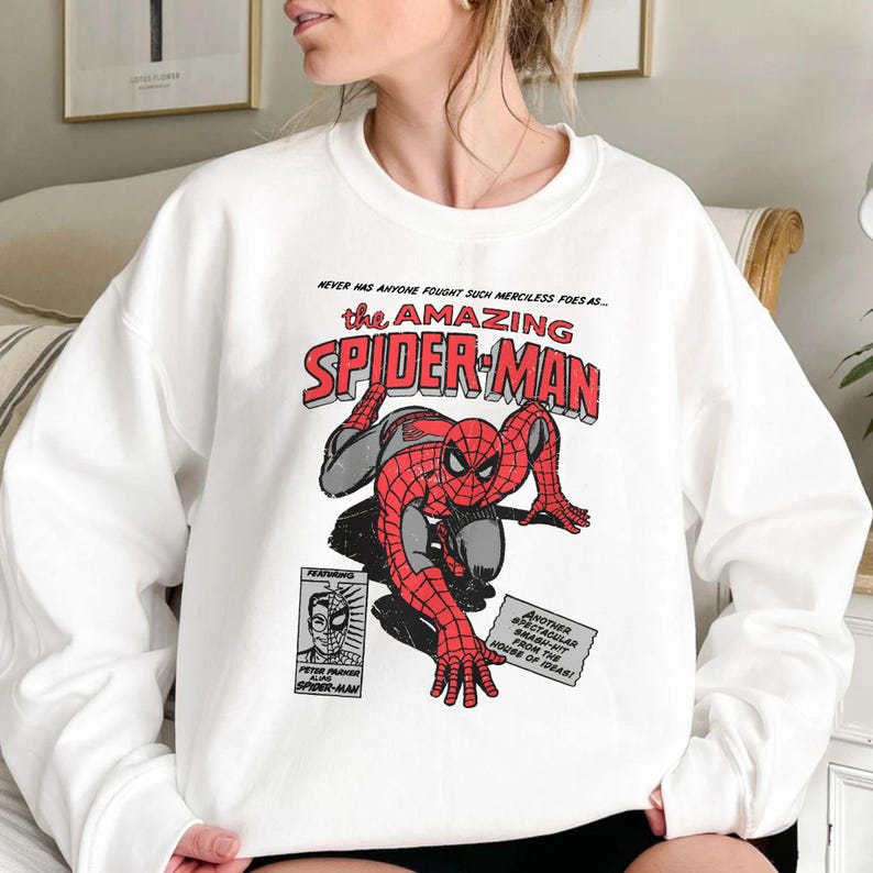 May include: White sweatshirt featuring a vintage comic book graphic of Spider-Man. The design includes the text "the AMAZING SPIDER-MAN" in red and black, with additional comic book elements.
