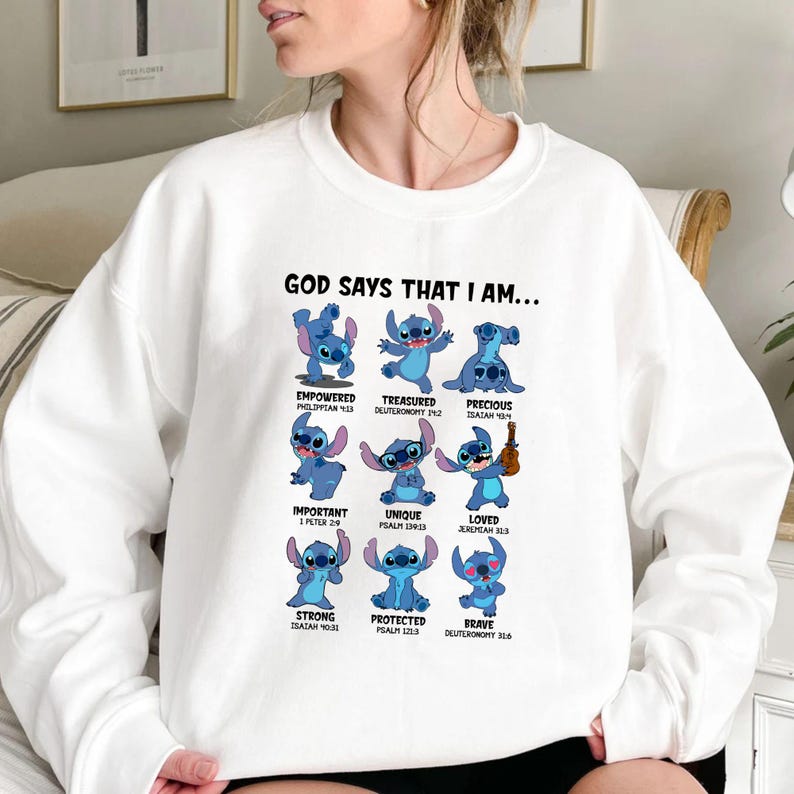 Op de afbeelding: Witte sweatshirt met een afbeelding van het personage Stitch uit Lilo & Stitch. Het ontwerp bevat Stitch in verschillende poses met de woorden "GOD SAYS THAT I AM..." en inspirerende woorden als "Empowered" en "Loved".