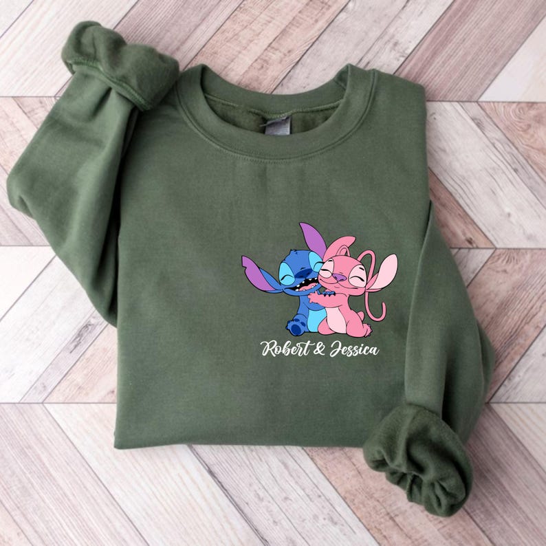 May include: An olive green sweatshirt with a graphic of two cartoon characters hugging, one blue and one pink. The text "Robert & Jessica" is printed below. The sweatshirt has long sleeves.