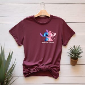 May include: A maroon t-shirt featuring a cartoon graphic of two embracing characters, one blue and one pink. The text "Robert & Jessica" is printed below the graphic. The shirt is displayed on a wooden hanger.