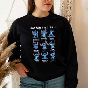 Op de afbeelding: Zwart sweatshirt met een afbeelding van Stitch en de tekst "GOD SAYS THAT I AM..." samen met positieve woorden als "Empowered", "Treasured" en "Precious". Het ontwerp bevat blauwe Stitch-illustraties.