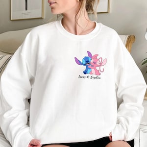 May include: White sweatshirt with a cartoon illustration of two embracing characters, one blue and one pink. The text "Lucas & Sophia" is printed beneath the characters in a cursive font. The sweatshirt is a casual style.