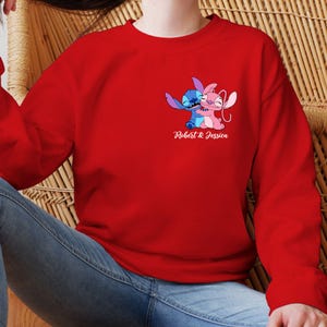 May include: A red sweatshirt with a graphic of two cartoon characters, one blue and one pink, in an embrace. The text "Robert & Jessica" is printed below the graphic in white script. The sweatshirt is made of a soft material.