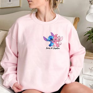 May include: Pale pink sweatshirt with a cartoon illustration of two characters, one blue and one pink, in an embrace. The text "Lucas & Sophia" is printed below the graphic in black script. The sweatshirt has a round neck.