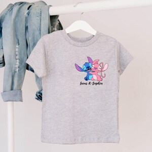 May include: A gray t-shirt featuring a cartoon image of two characters embracing, one blue and one pink. The text "Lucas & Sophia" is printed below the image. The shirt is hanging on a white hanger.
