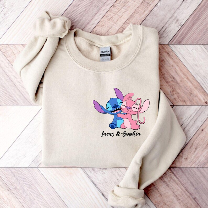 May include: A beige sweatshirt with a graphic of two cartoon characters embracing, one blue and one pink. The text "Lucas & Sophia" is printed beneath the graphic. The sweatshirt is folded on a wooden surface.